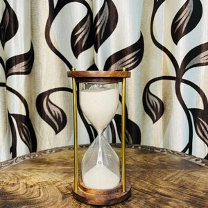 Könnte beinhalten: Eine hölzerne Sanduhr mit Messingstützen und einer durchsichtigen Glaskammer, die mit weißem Sand gefüllt ist. Die Sanduhr steht auf einer Holzoberfläche, mit einem gemusterten Vorhang im Hintergrund.