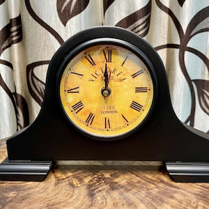 May include: Black wooden mantel clock with a round face featuring Roman numerals and the words "Victoria Station London 1747". The clock has a curved top and a rectangular base. The clock face is yellow.