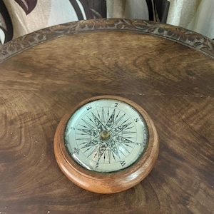 Puede incluir: Una brújula vintage con base de madera. La esfera de la brújula presenta un diseño detallado de estrella con marcas direccionales. La brújula está sobre una superficie de madera, lo que sugiere un tema náutico o de exploración.