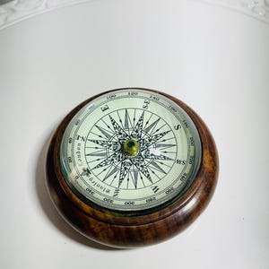 May include: A round magnetic compass with a polished dark brown wooden casing and a glass top. The cream-coloured dial features black cardinal directions, degree markings, and the text Stanley London printed in a decorative script font.