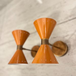 Op de afbeelding: Twee oranje wandkandelaars met een kegelvormig ontwerp en messing details. Elke kandelaar heeft kleine gaatjes aan de onderrand. De kandelaars zijn bevestigd aan een muur met een marmerpatroon.
