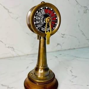 May include: A vintage brass engine room telegraph with a wooden base. The circular dial displays speed commands like "Full," "Half," and "Slow," with "Stop" in red. A gold-colored key hangs below the dial, adding to its nautical aesthetic.