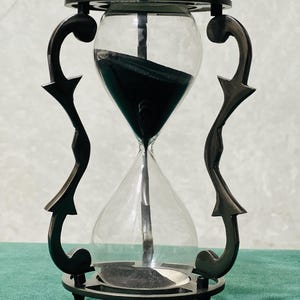 May include: An hourglass with a dark metal frame featuring ornate, curved side supports and a circular base. The clear glass bulbs contain fine black sand, with a portion currently in the top bulb and a small amount settled in the bottom bulb.