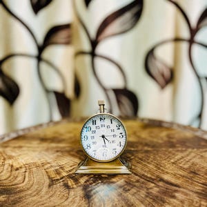 May include: A small, antique-style brass clock with a white face and black numerals. The clock sits on a decorative brass base and displays the time. The background features a wooden surface and a patterned curtain.