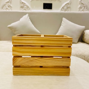 May include: A natural wood organizer with a square shape and a divided interior. The wooden crate is constructed with horizontal slats, showcasing the wood grain. The organizer is designed to hold items and is placed on a white surface.