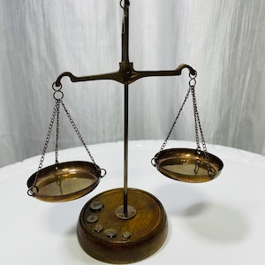 Peut inclure: Une balance en laiton composée de deux plateaux suspendus par de fines chaînes à une barre horizontale centrale. La balance est fixée sur une tige verticale montée sur un socle circulaire en bois, incluant plusieurs petits poids en laiton.