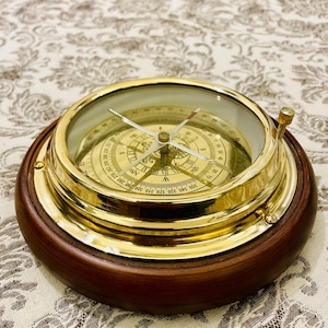 Puede incluir: Una brújula de latón vintage con tapa de cristal, colocada en una base de madera oscura. La esfera de la brújula presenta marcas direccionales y una aguja. La carcasa de latón tiene un acabado pulido y la base de madera es de color marrón oscuro. Un instrumento náutico clásico.