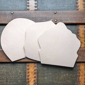 May include: Two blank, cream-coloured, wooden shapes, one round and one with a scalloped edge, are placed on top of a vintage wooden trunk with brass accents.