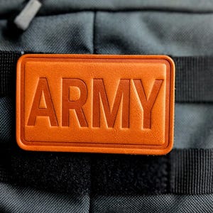 May include: An orange leather patch with the word "ARMY" embossed in capital letters. The patch is rectangular with rounded corners and is attached to a gray backpack with black straps.