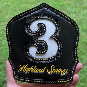 May include: A black leather shield-shaped fire helmet front featuring a large white number 3 with a black border. The text Highland Springs is embossed in yellow at the bottom. The entire piece is accented with yellow stitching along the edges.
