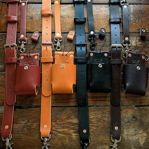 May include: A collection of leather straps in various colors, including brown, tan, and black, with metal hardware. Each strap has a small pouch attached. The straps are arranged vertically against a wooden background.