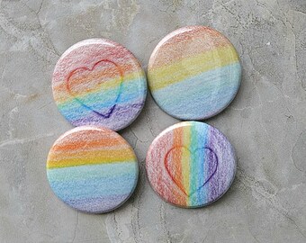Puede incluir: Cuatro botones redondos con diseños de lápices de colores arcoíris. Dos botones presentan un contorno de corazón sobre un degradado arcoíris, mientras que los otros dos tienen un degradado arcoíris. Los botones están dispuestos sobre una superficie gris.