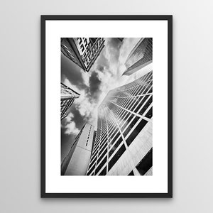 Op de afbeelding: Zwart-witfoto van hoge gebouwen in een stad, gezien vanaf de onderkant, met een bewolkte lucht erboven.