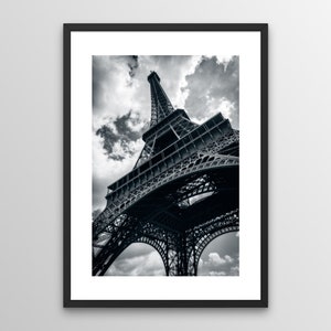 Op de afbeelding: Zwart-witfoto van de Eiffeltoren in Parijs, Frankrijk, in een zwarte lijst. De smeedijzeren tralietoren wordt van onderaf bekeken, tegen een achtergrond van een bewolkte lucht.