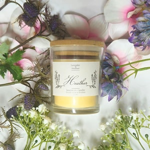 May include: A glass jar candle with a wooden lid, labeled "Heather" with floral accents, surrounded by purple and white flowers. The candle has a yellow wax interior. The arrangement is set against a white background.