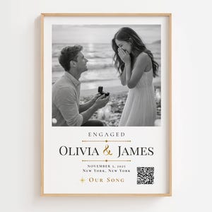 May include: A framed vertical print featuring a black and white photograph of a marriage proposal on a beach. Below the photo, the text reads ENGAGED, OLIVIA & JAMES, NOVEMBER 5, 2025, NEW YORK, NEW YORK, and OUR SONG, accompanied by a QR code.