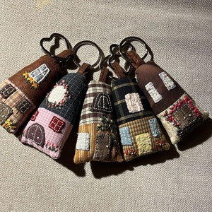 Può includere: Cinque portachiavi in ​​tessuto a forma di casa con diversi motivi e colori. I portachiavi sono marroni, rosa, gialli, a quadri e marroni con fiori rossi.