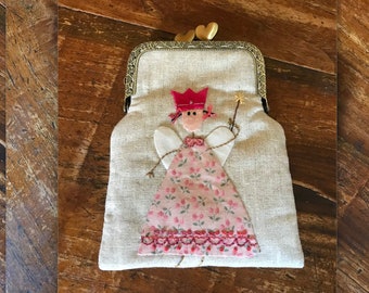 May include: A small, beige fabric coin purse with a gold clasp. The purse features an embroidered image of a fairy with a pink dress, white wings, and a red crown. The fairy is holding a wand with a star on the end.