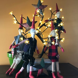 Op de afbeelding: Vijf handgemaakte stoffen gnomen met rode mutsen en witte gezichten zijn gerangschikt rond een tak met witte lampjes en gele bloemen. De gnomen dragen geruite en gestreepte kleding.