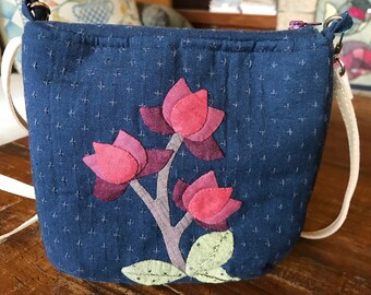 Op de afbeelding: Een klein blauw stoffen tasje met een witte band en een bloemenmotief. De bloemen zijn roze, paars en groen en zijn op de tas geplakt.