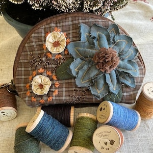 May include: A brown and tan plaid fabric pouch with a zipper closure. The pouch is decorated with embroidered flowers and a blue fabric flower. The pouch is surrounded by spools of yarn in various colours, including brown, green, blue, and tan. The spools are labelled "Rustic Wool Moire".
