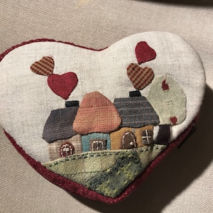 Puede incluir: Una bolsa de tela en forma de corazón con un borde rojo. La bolsa presenta un diseño de patchwork de casas con corazones rojos y un paisaje verde y marrón.