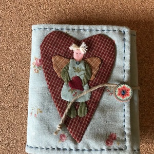 Puede incluir: Un diario de tela hecho a mano con una cubierta azul claro, con un aplique en forma de coraz&oacute;n con un dise&ntilde;o de &aacute;ngel cosido. El &aacute;ngel sostiene un coraz&oacute;n, y el diario se asegura con un bot&oacute;n y un cierre de cuerda. Acentos florales est&aacute;n dispersos en la cubierta.