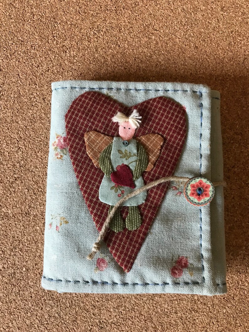 Puede incluir: Un diario de tela hecho a mano con una cubierta azul claro, con un aplique en forma de coraz&oacute;n con un dise&ntilde;o de &aacute;ngel cosido. El &aacute;ngel sostiene un coraz&oacute;n, y el diario se asegura con un bot&oacute;n y un cierre de cuerda. Acentos florales est&aacute;n dispersos en la cubierta.