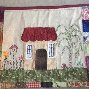 May include: A beige fabric zippered pouch with a quilted design featuring a house with a red roof, a tree, and a birdhouse. The house has two windows and a brown door. The pouch is surrounded by a red border.