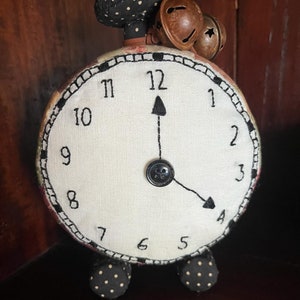 Op de afbeelding: Een witte stoffen klok met zwarte cijfers en wijzers, een zwarte en witte stippenstof bovenop en drie roestige bellen bovenop. De klok staat op een donkerbruin oppervlak.