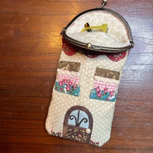 Può includere: Un'eccentrica custodia per occhiali a forma di casa, con un motivo a pois beige. La custodia presenta finestre applicate con dettagli floreali e una porta decorativa. L'interno, visibile, contiene un paio di occhiali gialli. La custodia è in tessuto e ha una chiusura in metallo.