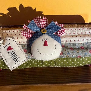 Può includere: Astuccio trapuntato con un volto sorridente di pupazzo di neve e un'etichetta con la scritta "I ❤️ Annie". L'astuccio è realizzato in tessuto patchwork rosso, bianco, blu e verde con motivi a stelle e fiori. Un fiocco rosso e blu adorna la parte superiore.