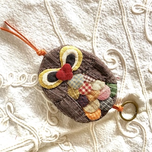 Op de afbeelding: Een bruine, uilvormige sleutelhanger van patchworkstof met een kleurrijk patchworkontwerp en een rood hart op de borst. De sleutelhanger heeft een oranje koord en een zilveren ring.