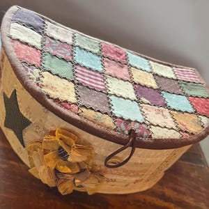 Könnte beinhalten: Eine handgefertigte, beige Stoffbox mit einem Patchwork-Deckel in verschiedenen Farben. Auf der Seite befindet sich ein dunkelgrüner Stern und eine gelbe Stoffblume. Die Box hat eine braune Umrandung und einen Leder-Schlaufenverschluss.