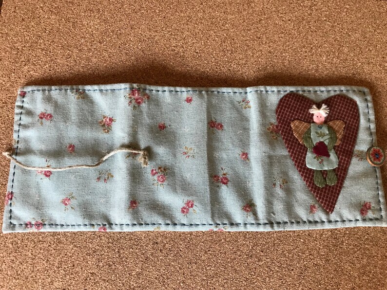 Puede incluir: Un organizador de tela azul claro con un estampado floral y costuras azul oscuro. Una aplicaci&oacute;n en forma de coraz&oacute;n con una figura de &aacute;ngel est&aacute; en el lado derecho. Tambi&eacute;n son visibles un bot&oacute;n y un cierre de cuerda.