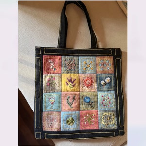 Puede incluir: Una bolsa de mano negra con un diseño de patchwork que presenta flores bordadas en varios colores. La bolsa tiene asas negras y un borde negro.