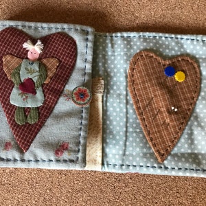 Puede incluir: Un libro de agujas de tela con una cubierta floral azul claro, adornado con una gran aplicaci&oacute;n de coraz&oacute;n granate y una peque&ntilde;a figura de &aacute;ngel. El interior es azul claro con lunares blancos y contiene un alfiletero de coraz&oacute;n.