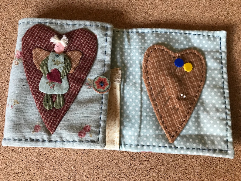 Puede incluir: Un libro de agujas de tela con una cubierta floral azul claro, adornado con una gran aplicaci&oacute;n de coraz&oacute;n granate y una peque&ntilde;a figura de &aacute;ngel. El interior es azul claro con lunares blancos y contiene un alfiletero de coraz&oacute;n.