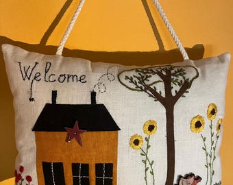 Puede incluir: Colgante de pared bordado con la palabra "Welcome" sobre una casa de color mostaza con tejado negro y una estrella roja. El diseño incluye un árbol, girasoles y un pajarito. El fondo es blanquecino.