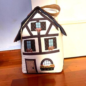 Puede incluir: Un tapón de puerta de tela blanco y marrón con forma de casa con una puerta marrón, ventanas y un techo marrón. La casa tiene una manija marrón en la parte superior.