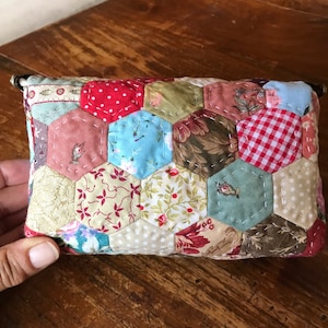 May include: A small, quilted pouch with a hexagonal pattern. The pouch is made of various fabrics in shades of red, green, blue, and white. The pouch has a zipper closure and two metal rings on the top.