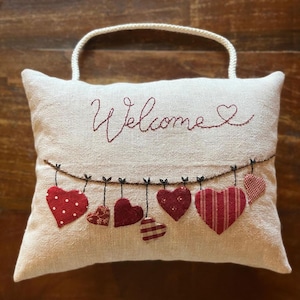 Può includere: Cuscino color crema con manico intrecciato, ricamato con la parola "Welcome" in corsivo rosso. Sotto, una fila di cuori appesi in vari motivi rossi aggiunge un tocco decorativo. Un affascinante oggetto di arredamento per la casa.