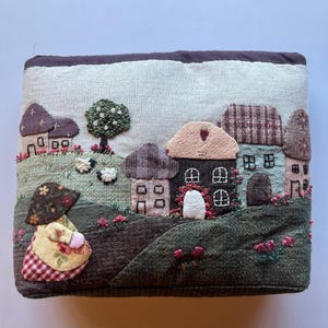 Könnte beinhalten: Eine beige Stofftasche mit einer gestickten Szene eines kleinen Dorfes mit Häusern, einem Baum und einer Frau in einem gelben Kleid und einem rot-weiß karierten Rock. Die Tasche ist mit braunem, grünem, rosa und rotem Stoff verziert.