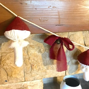 Peut inclure: Une guirlande de fête avec des champignons en tissu rouge et des nœuds bordeaux avec des cloches suspendus à une ficelle blanche.