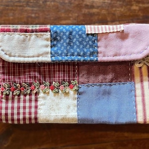 Könnte beinhalten: Patchwork-Clutch mit Stoffen in Rot, Blau und Weiß. Das Design umfasst Karomuster, Blumenmuster und Streifen, mit einer dekorativen floralen Bordüre. Der abgerundete Klappverschluss ist mit weißen Nähten befestigt. Ca. 20 cm breit.