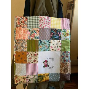 May include: A patchwork tote bag with a variety of floral and geometric patterns in shades of pink, green, yellow, and blue. The bag has a white canvas panel with a large embroidered letter "C" in the center.