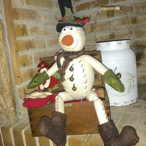 Può includere: Un pupazzo di neve bianco con un cappello a cilindro nero, guanti verdi e una sciarpa marrone è seduto su una scatola di legno. Il pupazzo di neve tiene una piccola bambola rossa e verde. Un bidone di latte bianco in metallo con un manico di legno è dietro il pupazzo di neve.