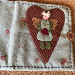 Puede incluir: Cartera de tela hecha a mano con un estampado floral y una aplicaci&oacute;n en forma de coraz&oacute;n con un &aacute;ngel. La cartera se asegura con un cierre de bot&oacute;n. El &aacute;ngel tiene alas y sostiene un coraz&oacute;n.