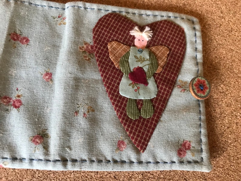 Puede incluir: Cartera de tela hecha a mano con un estampado floral y una aplicaci&oacute;n en forma de coraz&oacute;n con un &aacute;ngel. La cartera se asegura con un cierre de bot&oacute;n. El &aacute;ngel tiene alas y sostiene un coraz&oacute;n.