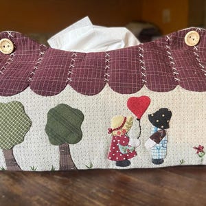 Op de afbeelding: Een decoratieve tissueboxhoes in de vorm van een huis, met een bordeauxrood dak en een crèmekleurig lichaam. De voorkant is versierd met geappliqueerde bomen en figuren van kinderen die een hartvormige ballon vasthouden. Houten knopen sieren het dak.